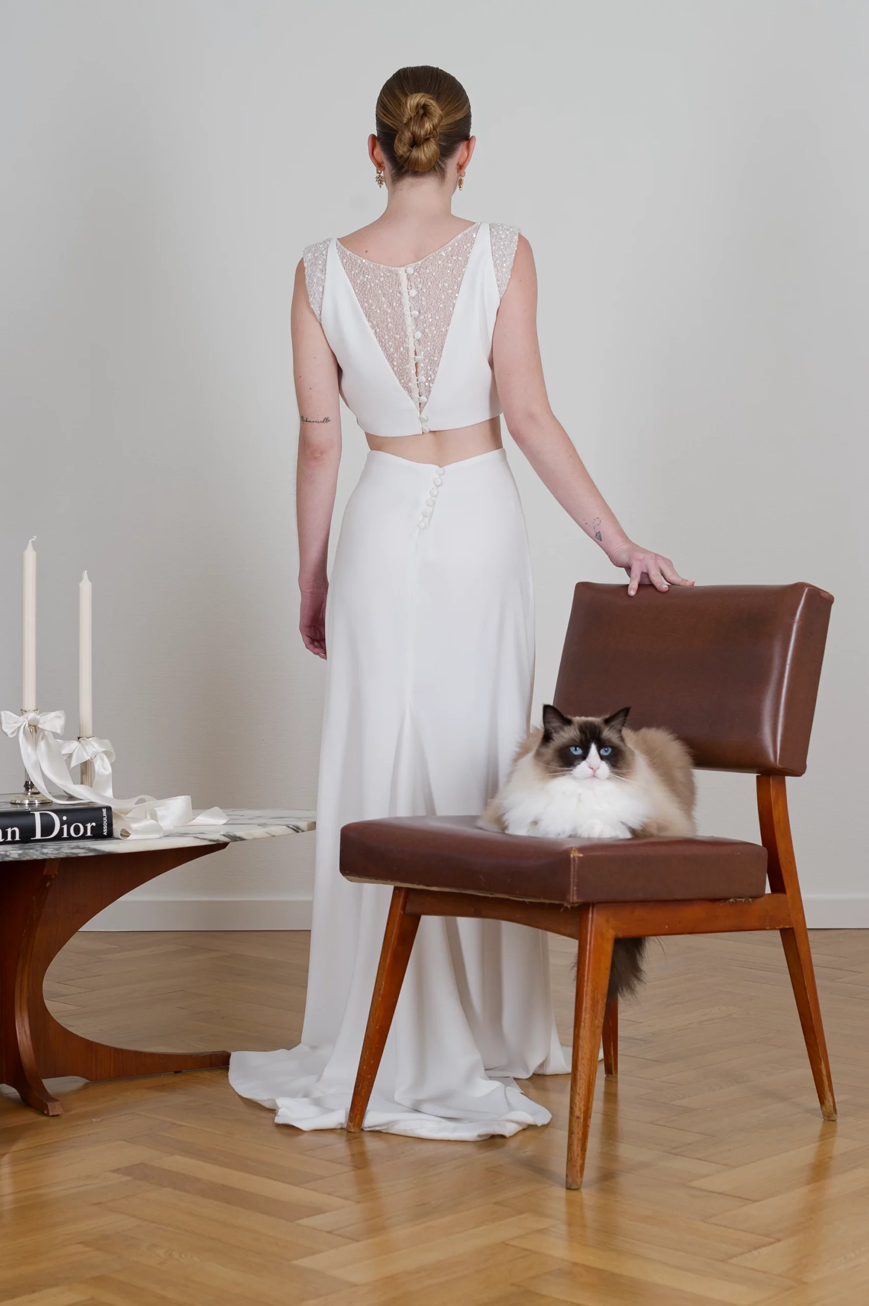 Mariée en robe élégante avec dos en dentelle, posant à côté d'un chat assis sur une chaise.