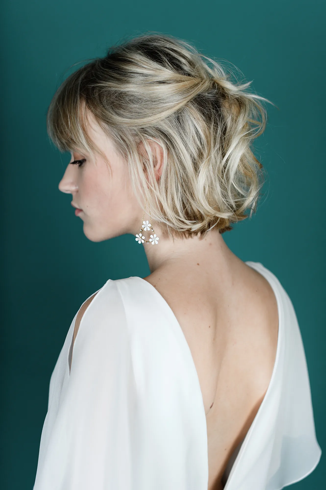 Vue arrière d'une femme portant une robe de mariée blanche fluide avec des manches transparentes et des boucles d'oreilles florales, avec un fond vert.