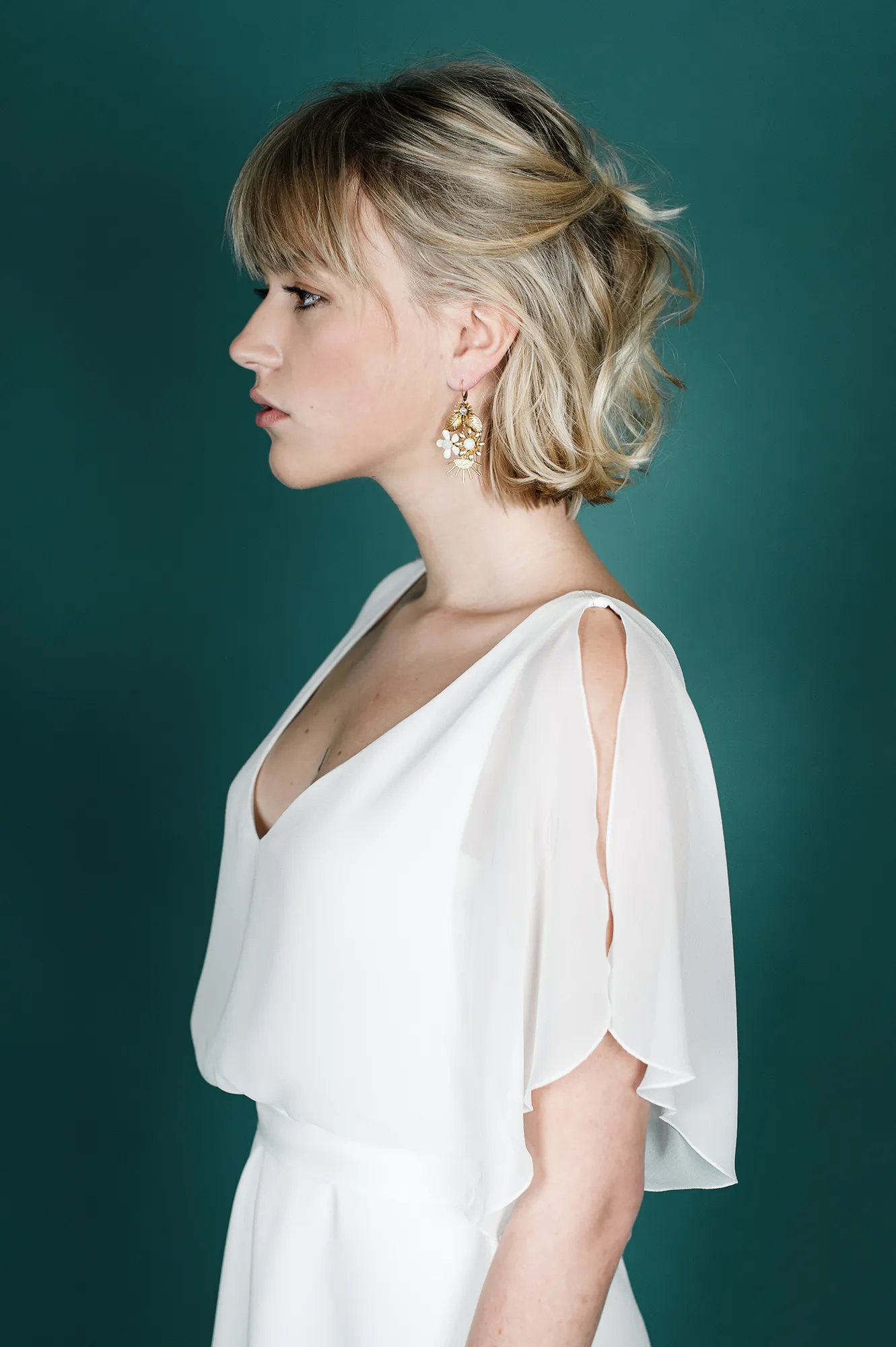 Femme portant une robe de mariée blanche fluide avec des manches transparentes et des boucles d'oreilles florales, vue de profil.