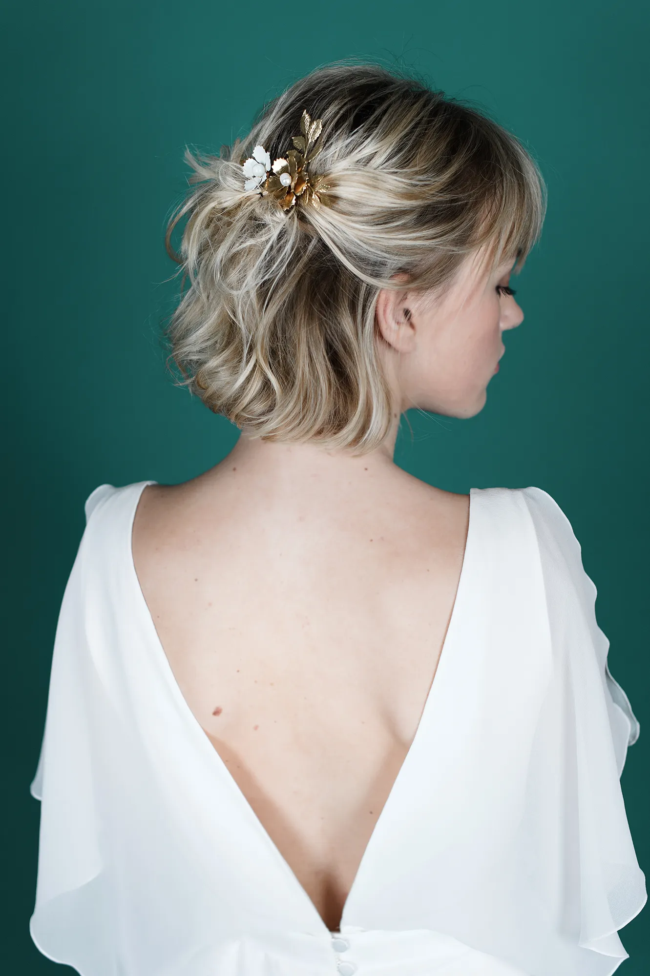 Vue arrière d'une femme portant la robe de mariée 'Chloé' avec un dos plongeant et des boutons décoratifs, et un bijou de cheveux floral doré.