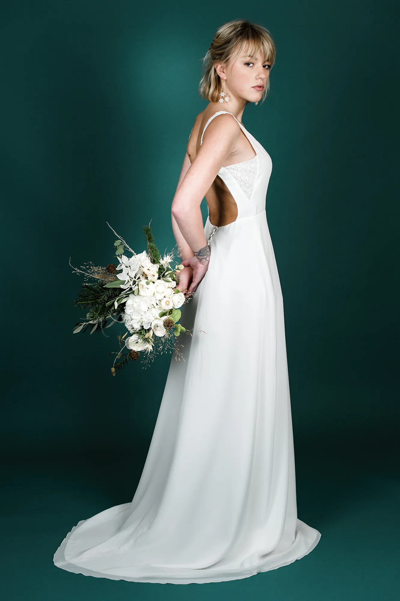 Femme portant la robe de mariée 'Garance', vue de profil, avec un dos ouvert, des détails en dentelle et tenant un bouquet de fleurs blanches.