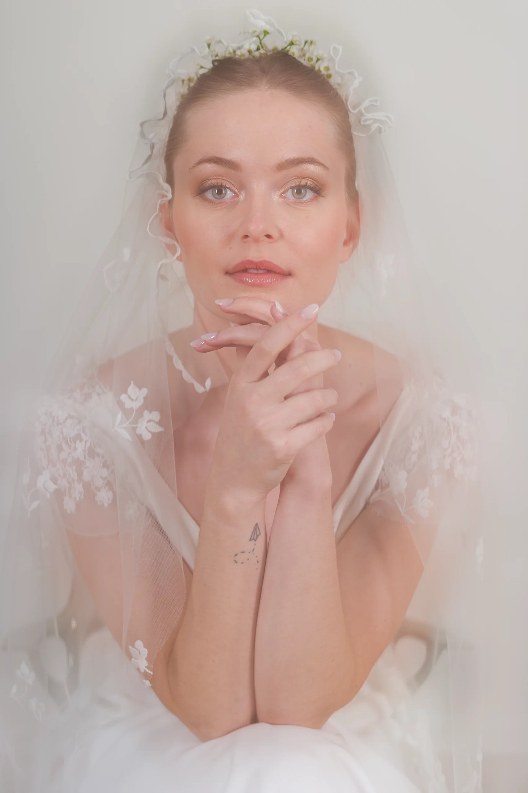 Portrait de la mariée avec un voile délicat et des manches en dentelle florale, les mains croisées près du visage pour une pose élégante.