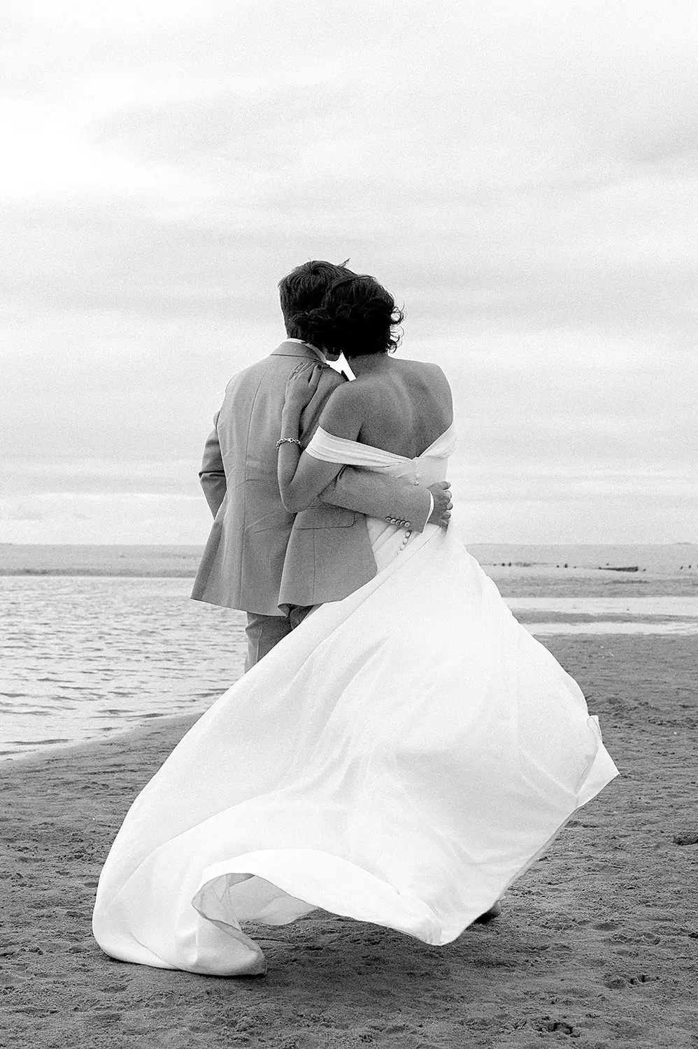 Jennifer - Robe de mariée en Dentelle de Calais – Création sur mesure Louise Dentelle à Toulouse Couple de mariés au bord de la mer, la mariée porte une robe Louise Dentelle épurée et fluide, dos nu, en mouvement sous le vent.