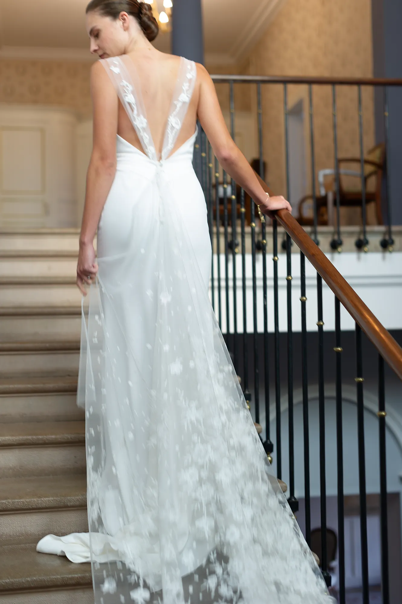 Robe de mariée Laura vue de dos, décolleté en V profond et traîne en tulle brodé, photographiée dans un escalier. Création Louise Dentelle à Toulouse.