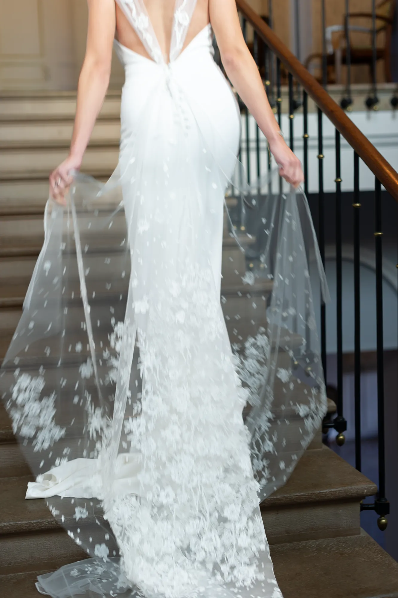 Robe de mariée Laura vue de dos, décolleté en V et traîne en tulle brodé en mouvement, tenue délicatement à la main. Création Louise Dentelle à Toulouse.