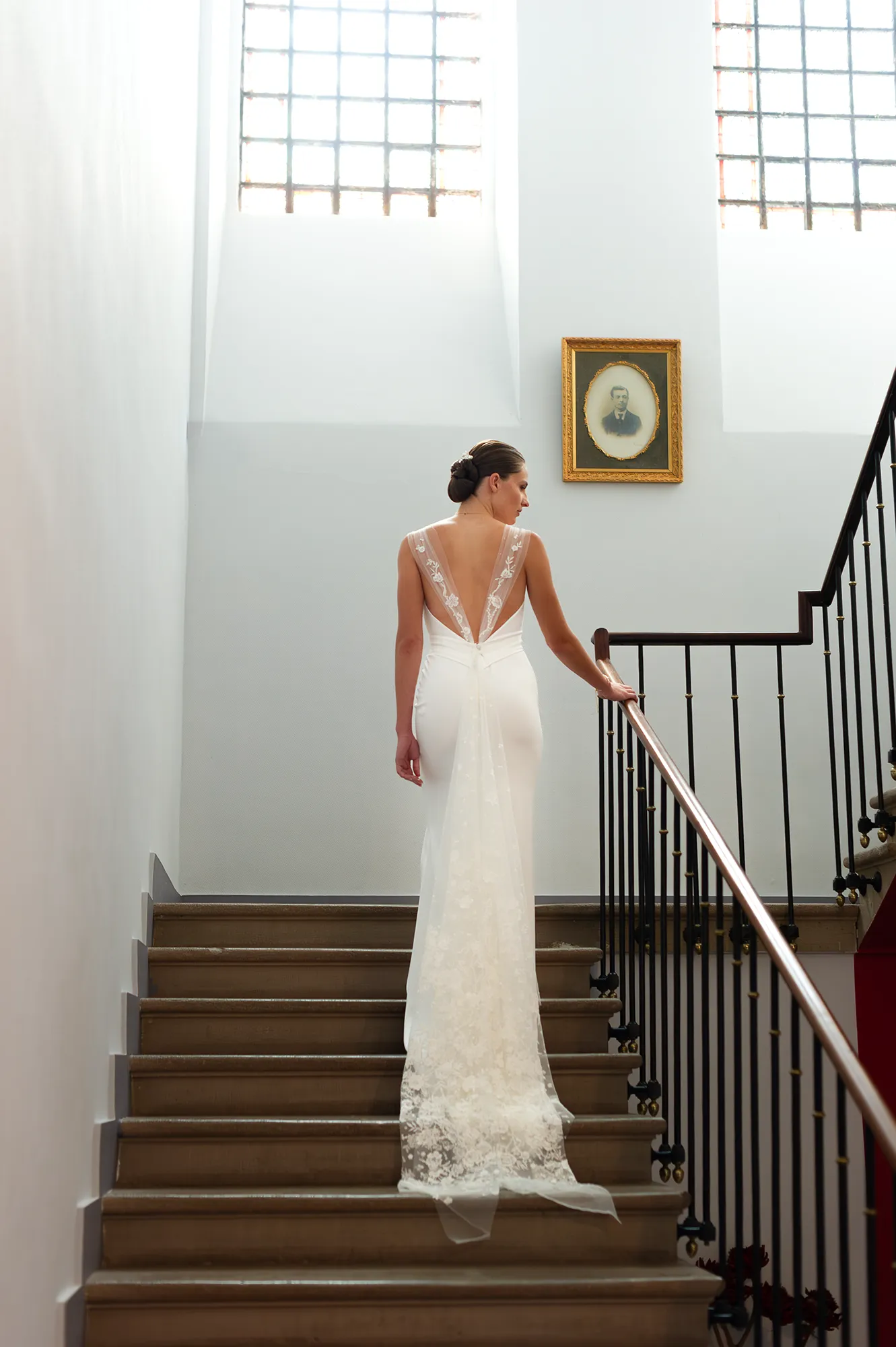Robe de mariée Laura vue de dos, décolleté en V profond et traîne en tulle brodé, photographiée dans un escalier baigné de lumière. Création Louise Dentelle à Toulouse.